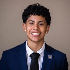 Rafael Farrera-Juarez smiles at the camera, with short dark curly hair, wearing a navy suit jacket, white shirt, black tie, and a round lapel pin, against a plain gray background.